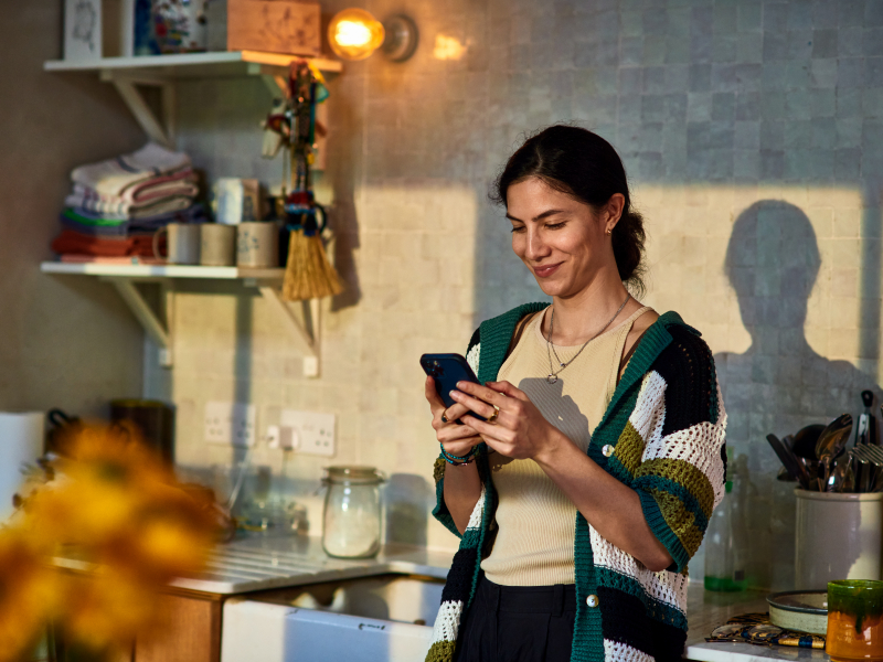 Lächelnde Frau steht in der Küche und schaut auf ihr Smartphone.